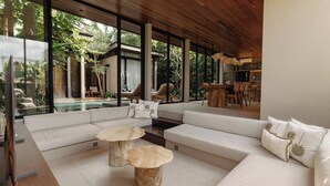 A modern living room with a large sectional sofa, two round wooden tables, and a dining area with wooden chairs. - The Akasha Canggu Luxury Villas (Canggu)