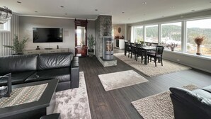 Een moderne woonkamer met een stenen openhaard, donker houten vloeren, een zwarte leren bank en een eethoek met uitzicht op de omgeving.