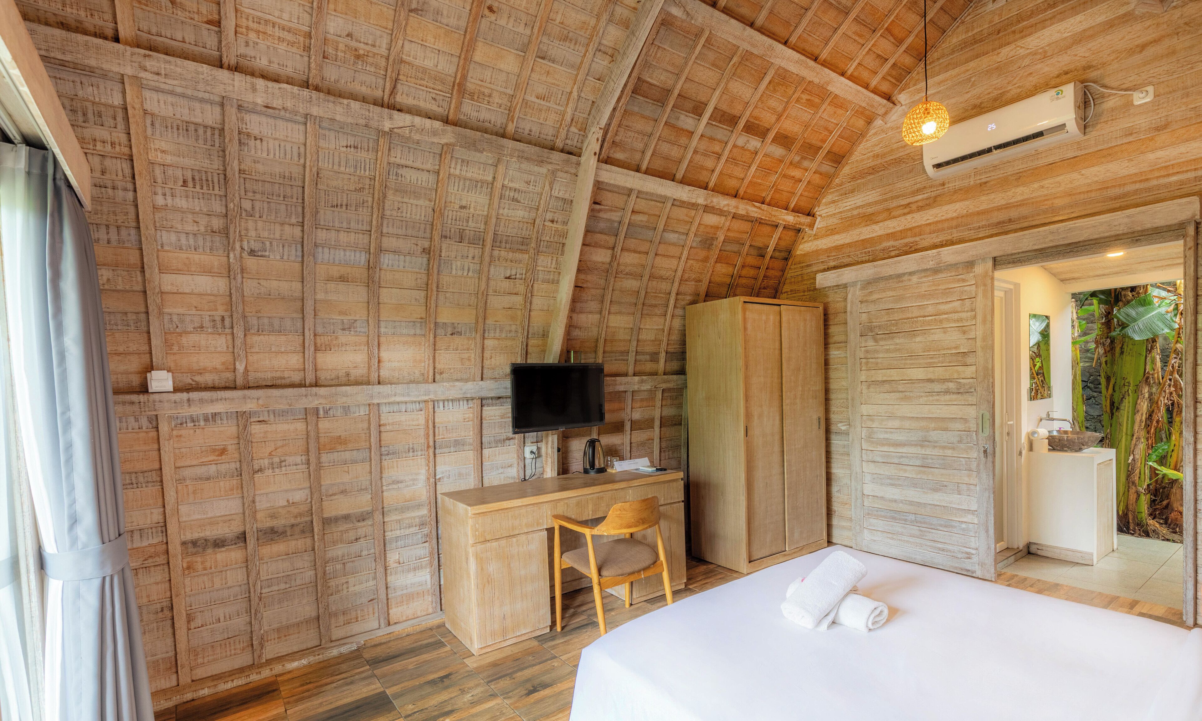 A bedroom with a sloped ceiling, wooden walls, a desk with a chair, a TV, a wardrobe, and a bathroom with a sink and shower.