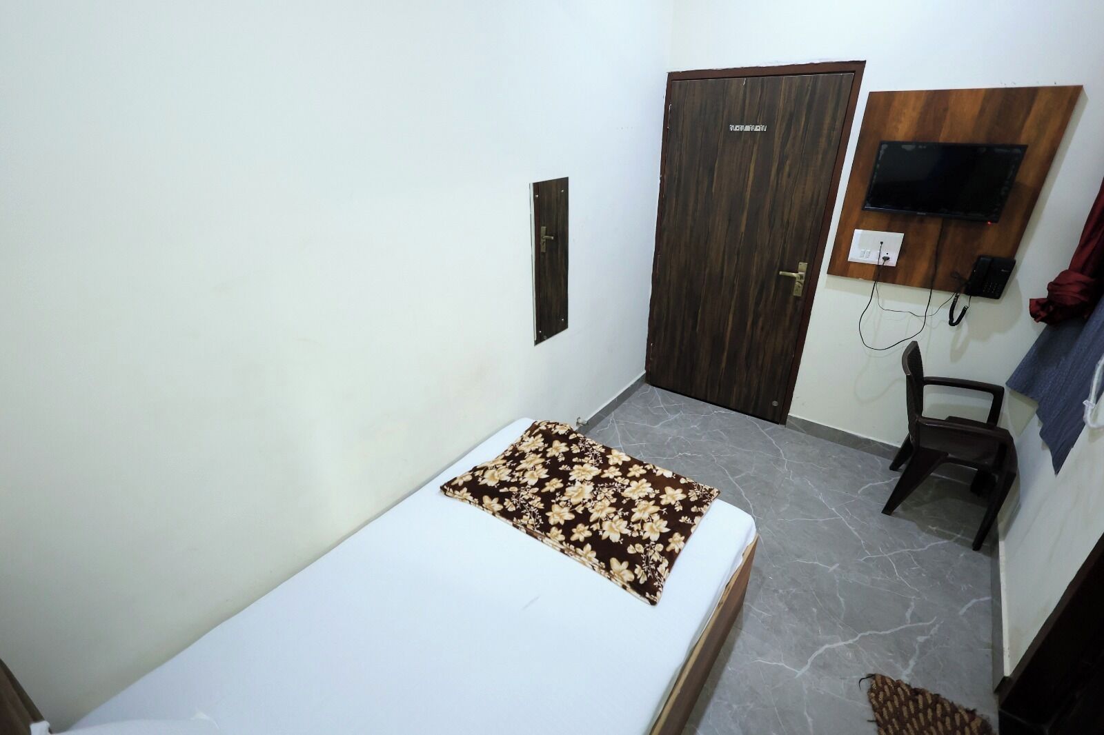 Una habitación pequeña y sencilla con una cama, un armario de madera, un televisor montado en la pared y una silla.
