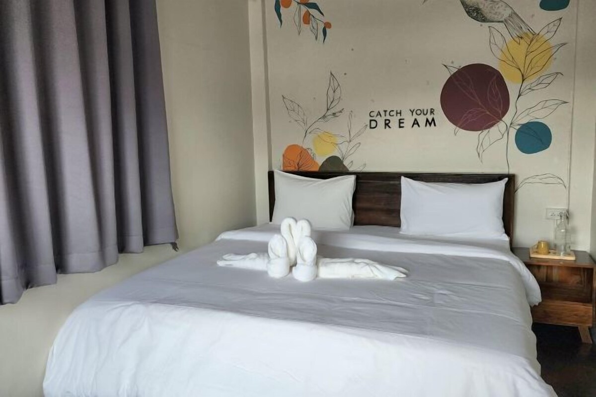 A neatly made bed with white linens and a towel arrangement, a wall with a motivational quote, and a simple bedside table with a glass.