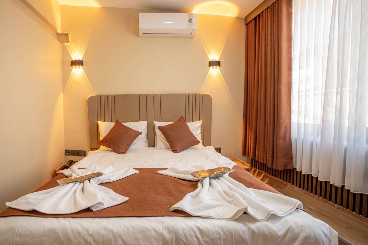 A hotel room with a large bed, two bedside tables, a wall-mounted air conditioner, and a window with curtains.