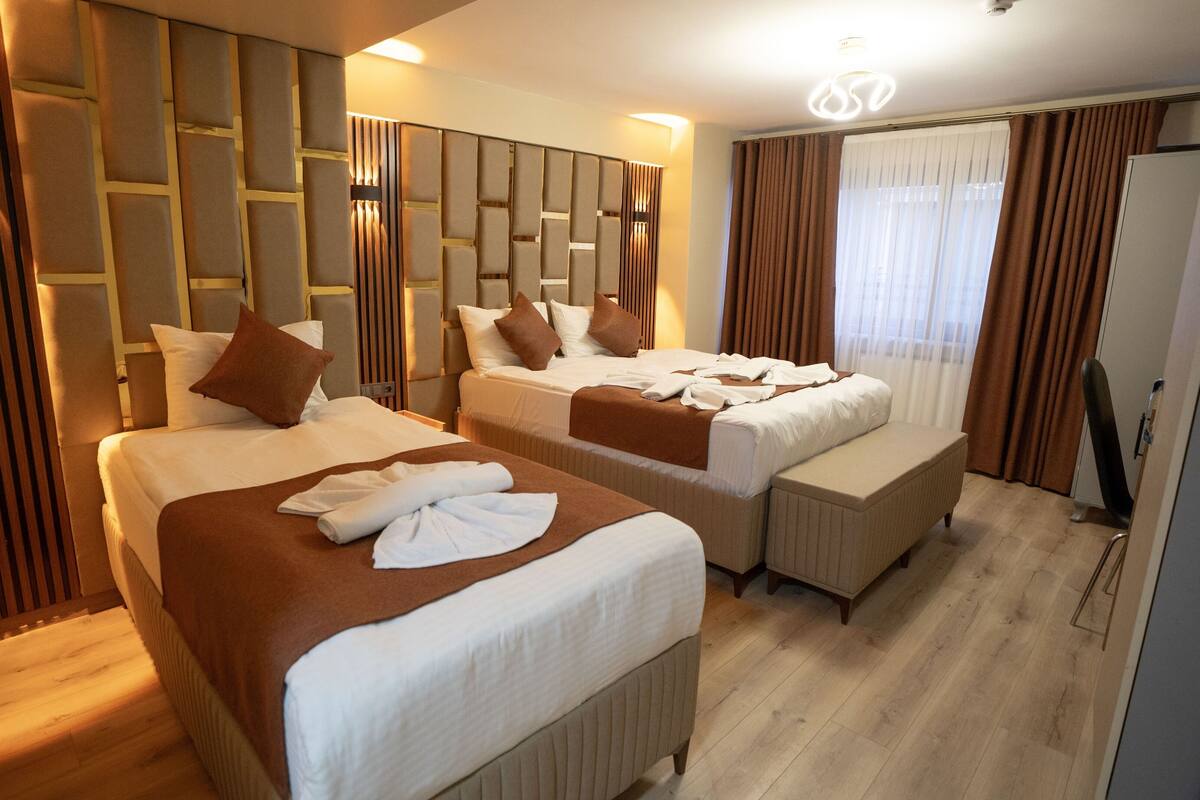 A hotel room with two beds, a wooden floor, a modern headboard, and a large window with curtains.
