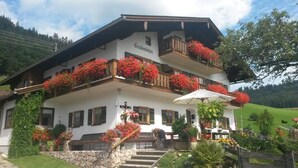 A traditional Alpine-style building with a balcony adorned with red flowers, a stone staircase, and a wooden fence.