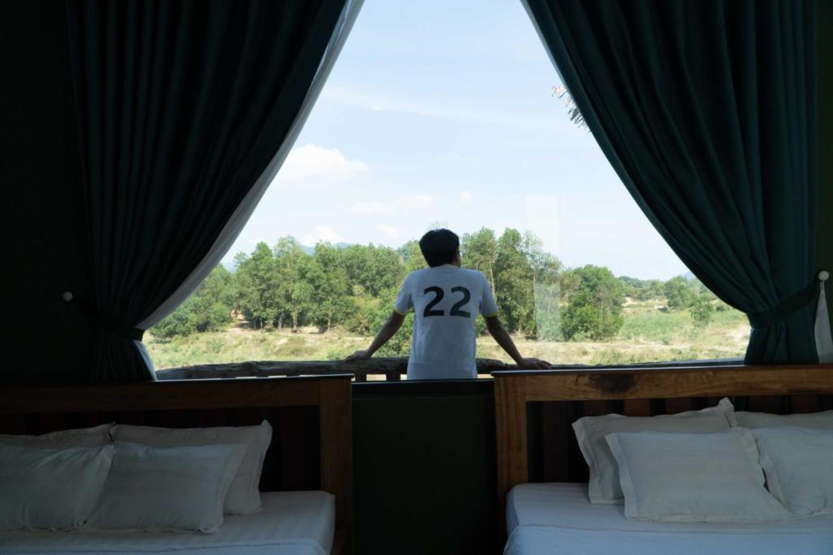 A person in a white shirt with the number 22 standing at a window with curtains, overlooking a natural landscape.