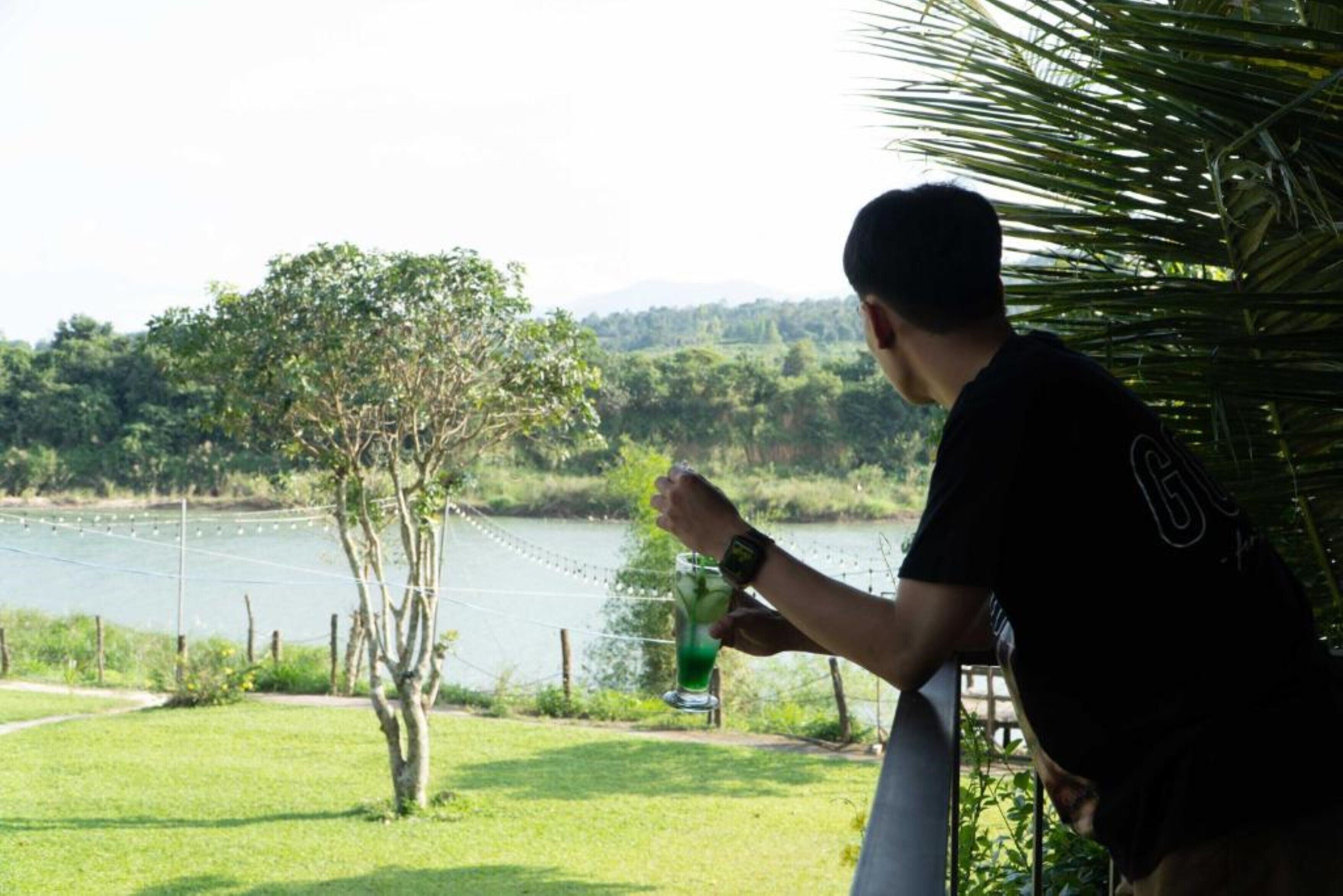 Une personne tenant une boisson près d’un lac, avec un arbre et une clôture au premier plan.
