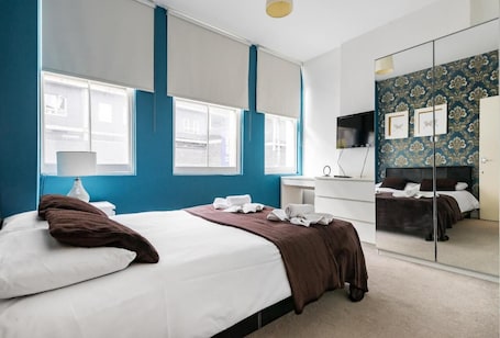 A modern hotel room with a large bed, a desk, a mirror, and a view of the city through the windows.