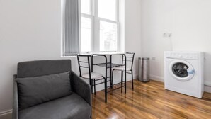 Una habitación con un sofá gris, una mesa de metal con sillas, una lavadora y una ventana con persianas.