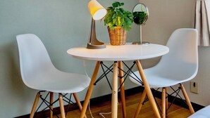 Une pièce comprenant une table ronde, deux chaises blanches, une lampe, une plante en pot, une horloge murale et un tableau encadré.