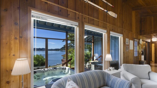 A wooden interior with a sofa, a table with a lamp, and a view of a hot tub and water body outside. - Captain s Retreat in Shelton (Shelton)