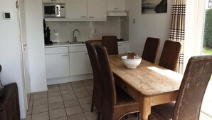 A compact kitchen with white cabinets, a microwave, and a coffee maker. A wooden dining table with wicker chairs is positioned in front of the kitchen. - Modern Beach Retreat – Sauna (Breskens)