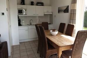 A compact kitchen with white cabinets, a microwave, and a coffee maker. A wooden dining table with wicker chairs is positioned in front of the kitchen. - Modern Beach Retreat – Sauna (Breskens)