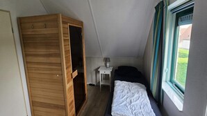 A bedroom with a bed, a wooden wardrobe, a bedside table with a lamp, and a window with curtains. - Modern Beach Retreat – Sauna (Breskens)