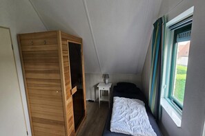 A bedroom with a bed, a wooden wardrobe, a bedside table with a lamp, and a window with curtains. - Modern Beach Retreat – Sauna (Breskens)