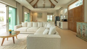 A spacious living area with a large sectional sofa, a wooden coffee table, and a kitchen with wooden cabinets and a large window. - Brand New Completed Chalong A  v124 (Chalong)