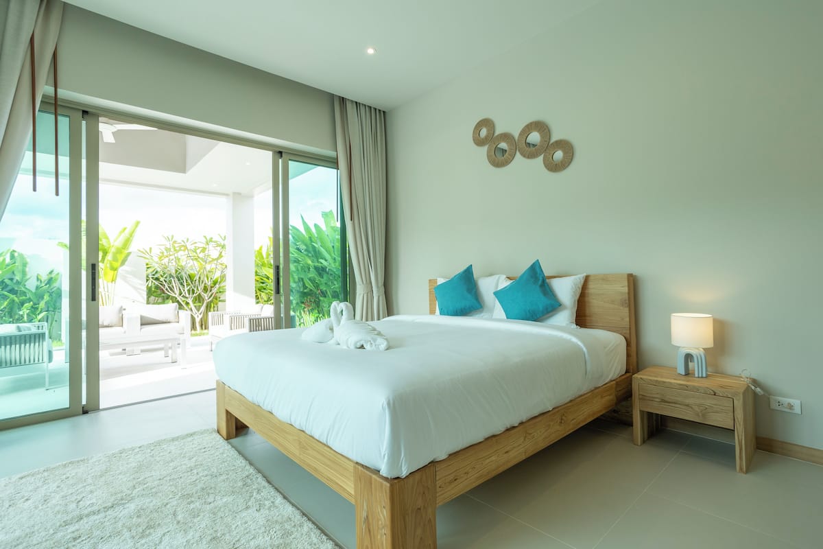 A spacious bedroom with a large bed, a nightstand, and a view of a lush garden through a sliding glass door.