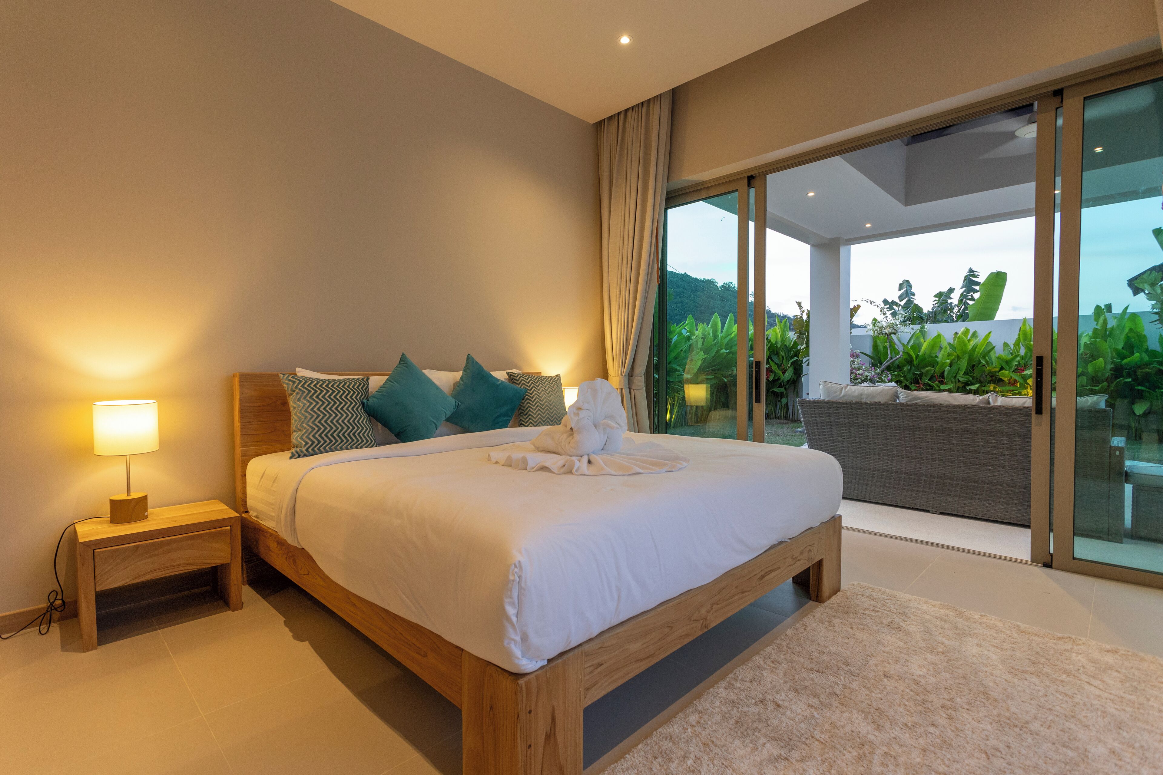 A bedroom with a wooden bed, a nightstand with a lamp, and a view of a balcony with plants.