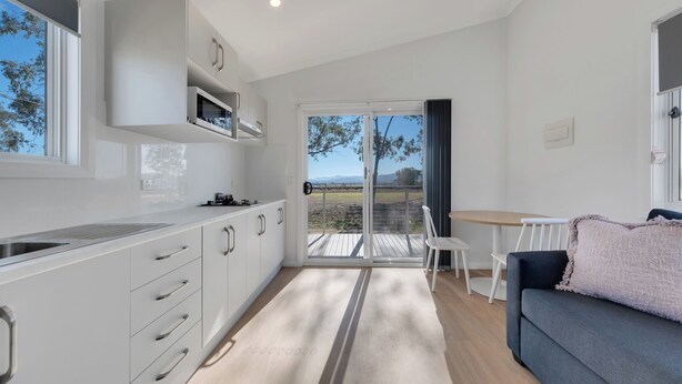 A modern kitchen with white cabinets and a large window, a cozy seating area with a dark sofa and a small table, and a sliding glass door leading to an outdoor deck. - Tamworth Holiday Park (Hillvue)