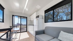 A modern kitchen with white cabinets and a built-in microwave, a grey sofa with a cushion, and a dining area with a table and chairs. - Tamworth Holiday Park (Hillvue)