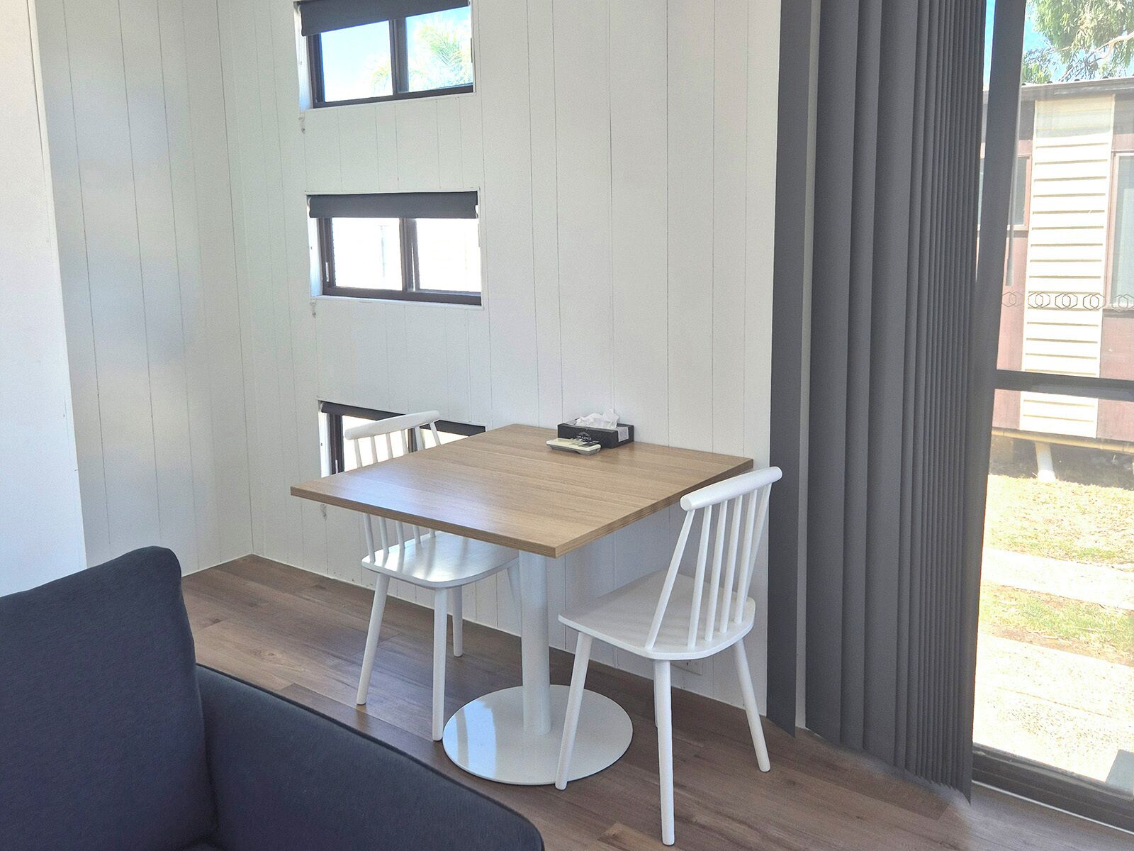 A room with a wooden table, two white chairs, a black sofa, and a window with curtains.