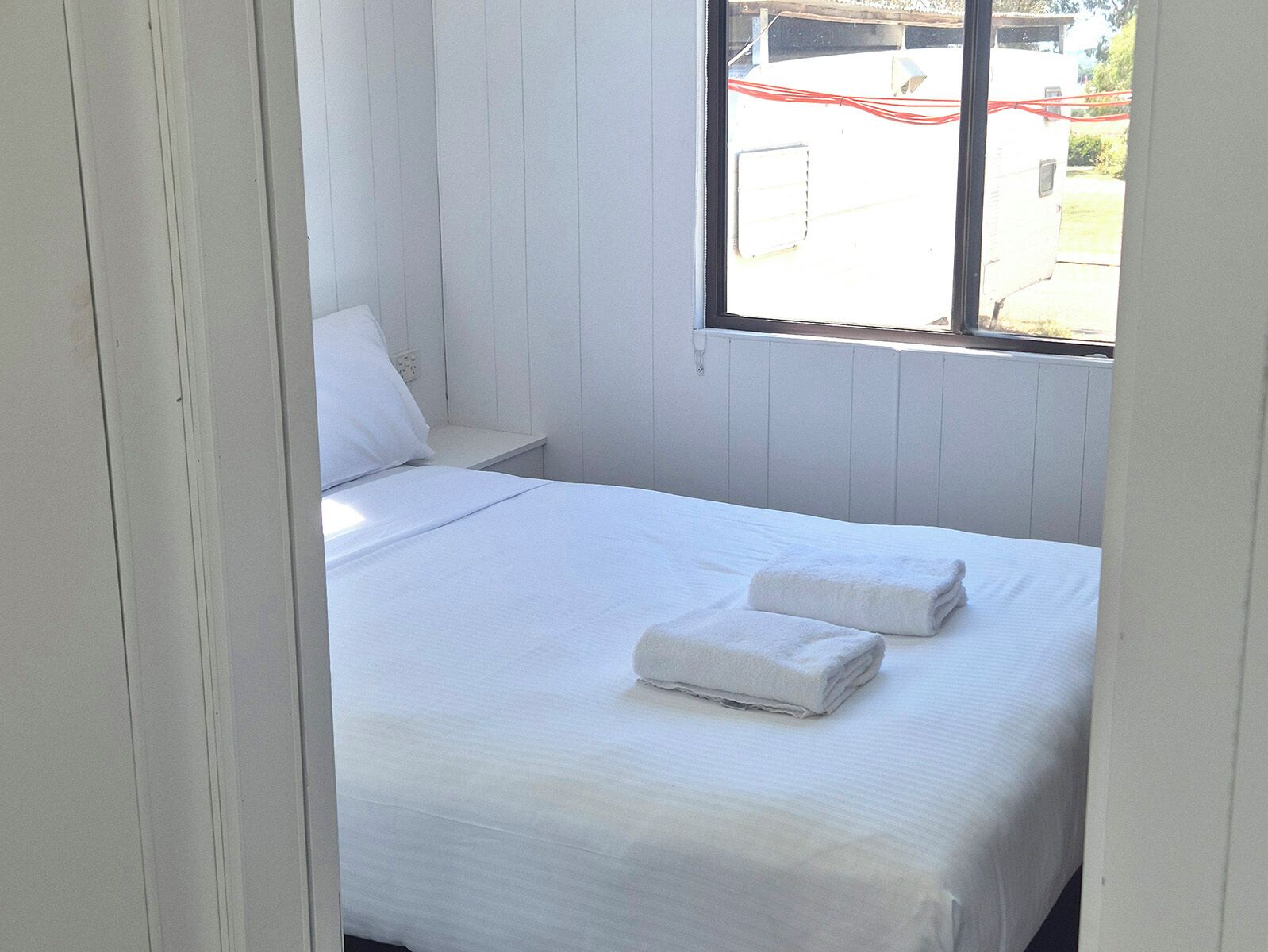 A single bed with white linens and two folded towels, a white nightstand, and a window with a view of the outside.