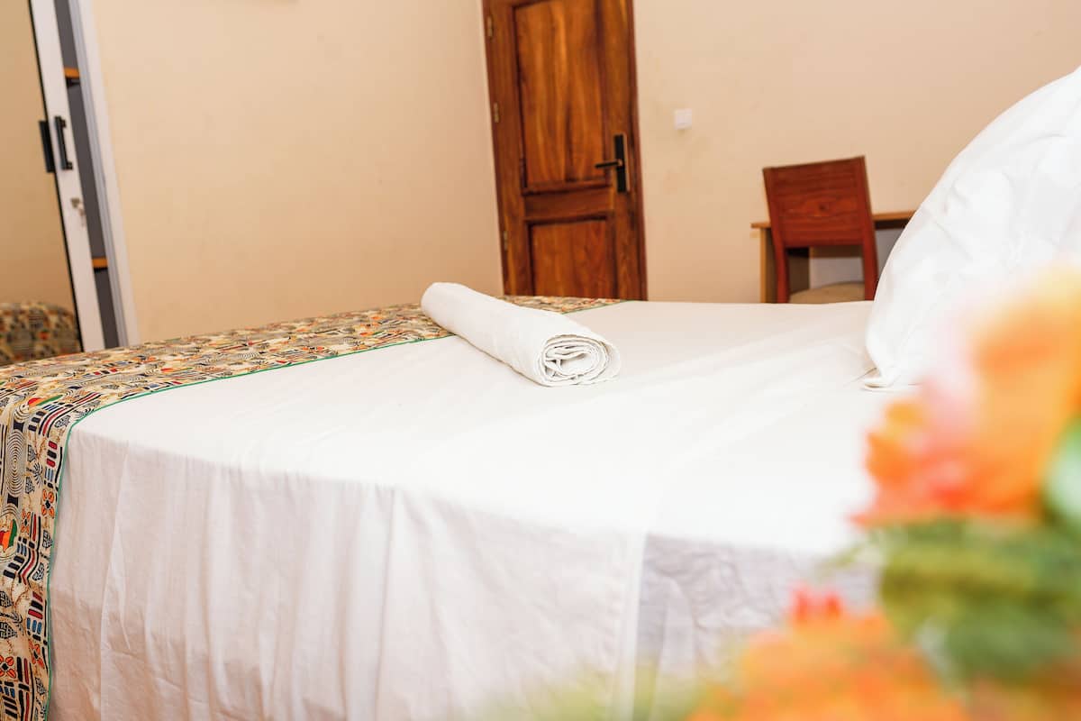 A neatly made bed with a rolled white towel, a wooden door in the background, and a chair to the right.