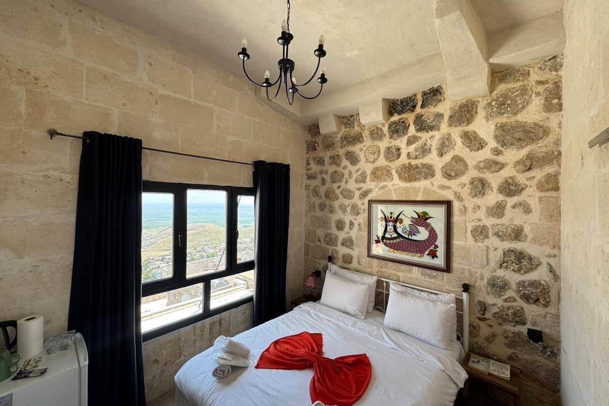 A room with a bed, a chandelier, a stone wall, a window with curtains, and a framed picture.