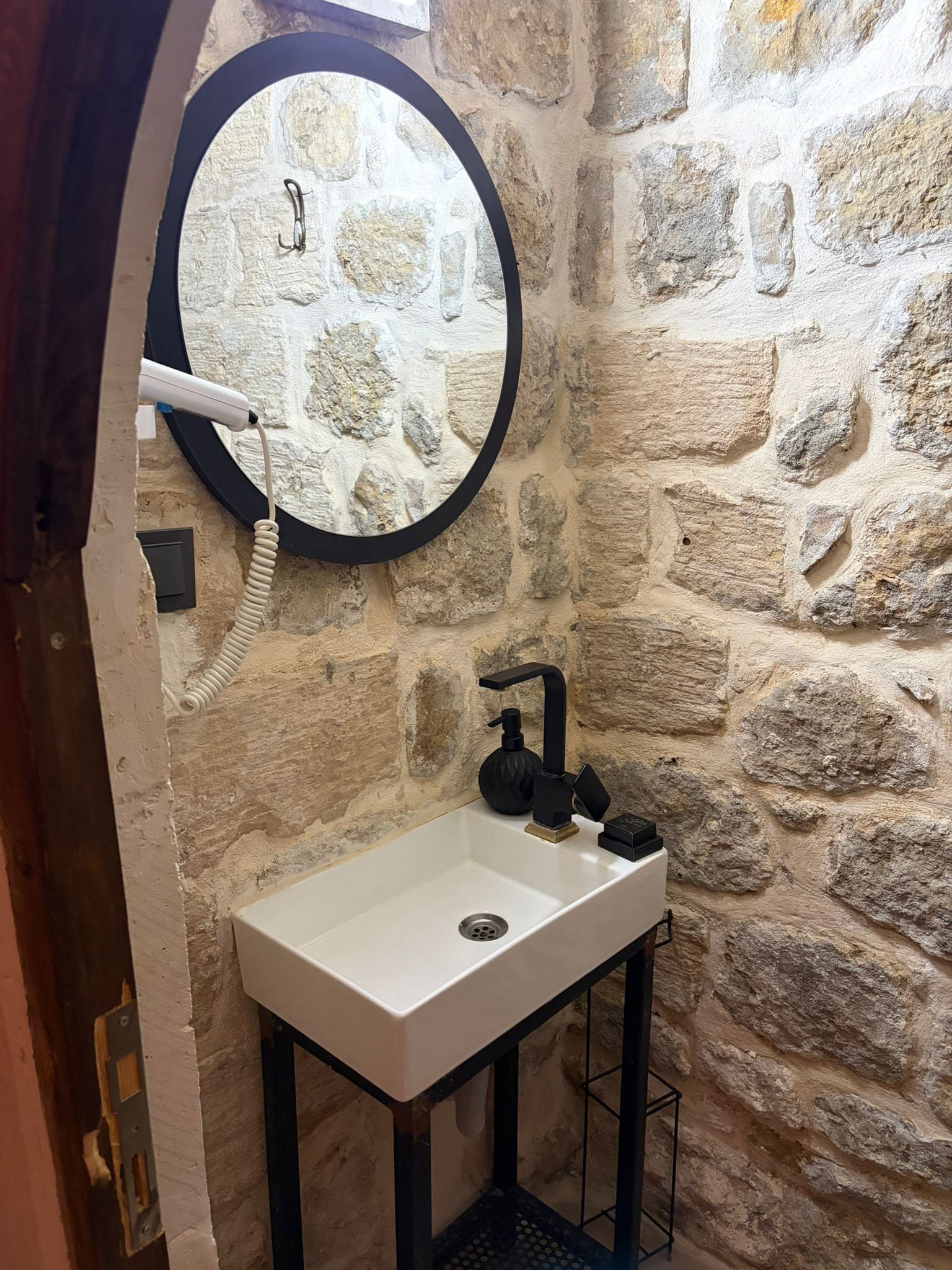 A bathroom with a stone wall, a round mirror, a white sink, and a black faucet.