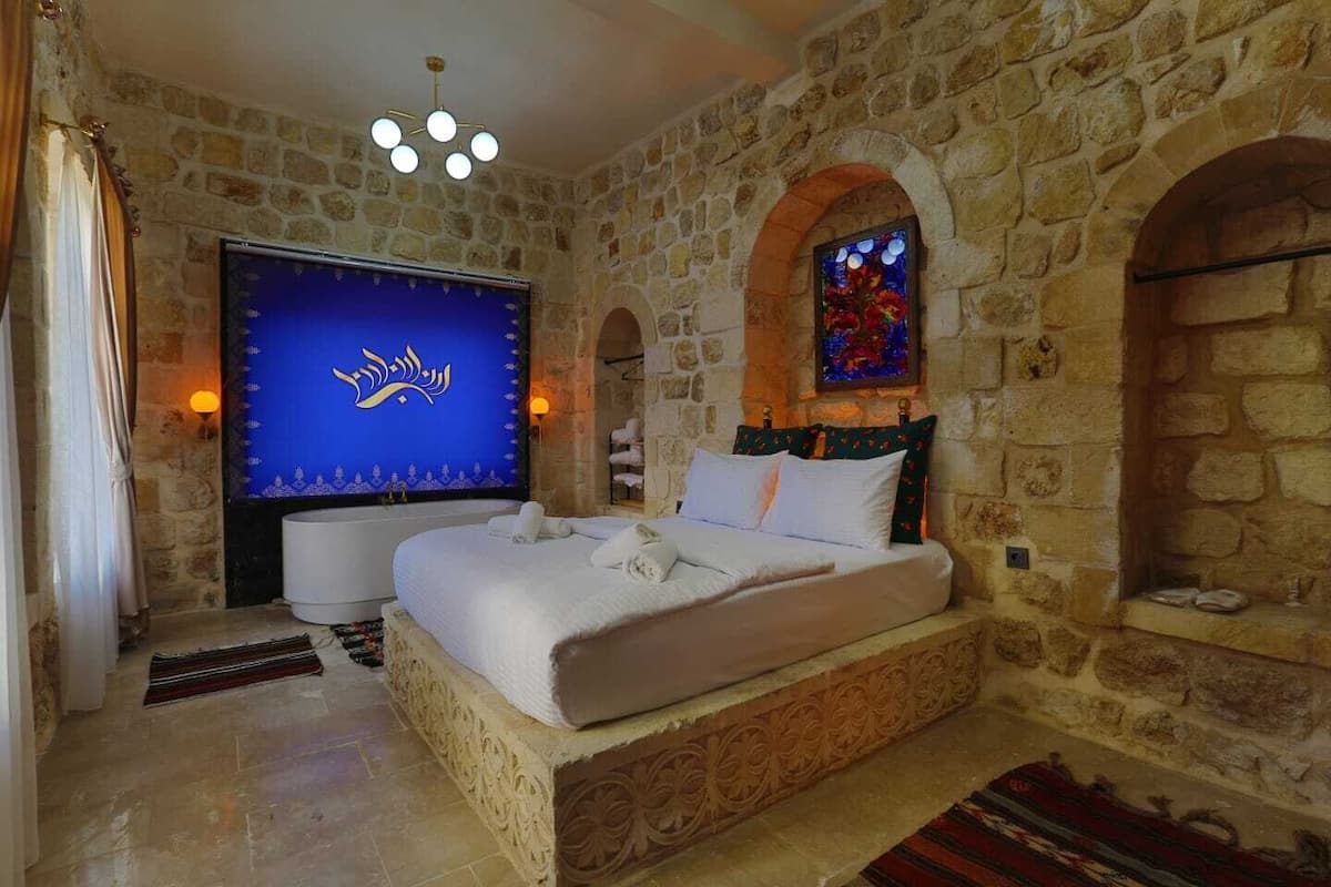 A bedroom with a stone wall, a bed with white linens, a large TV screen, and a decorative stained glass window.
