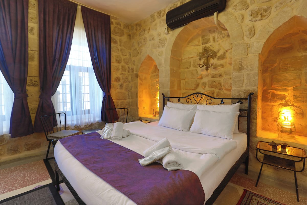 A bed with white linens, a purple throw blanket, and rolled towels. There is a chair and a small table with a lamp. The room has stone walls and arched doorways.