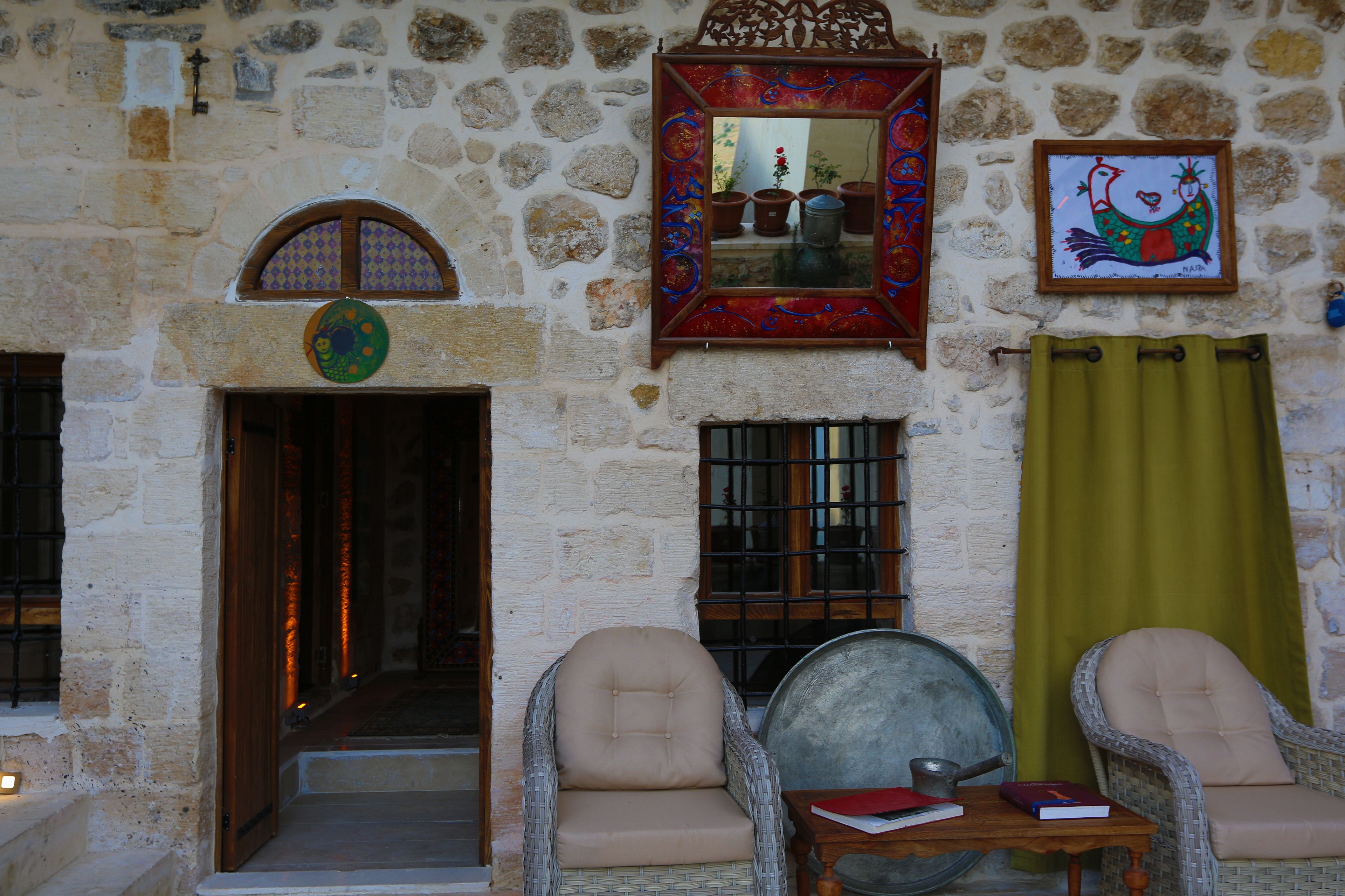 Una habitación con paredes de piedra, una puerta de madera, dos sillas de mimbre, una pequeña mesa con libros y un espejo con marco de colores.
