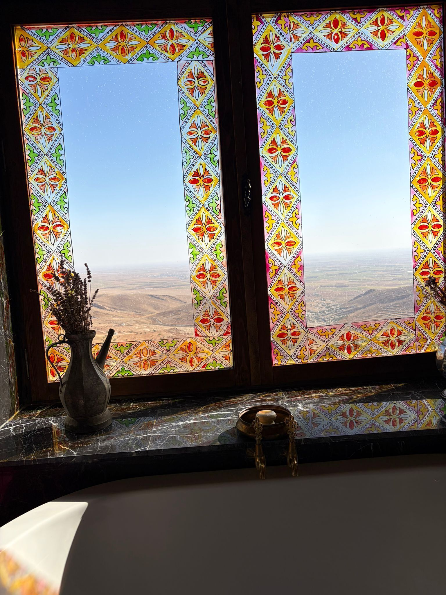 Ein Zimmer mit Blick durch ein Fenster mit farbigem Buntglas, einer Vase mit Trockenblumen und einem kleinen Tisch mit Kerzenhalter.