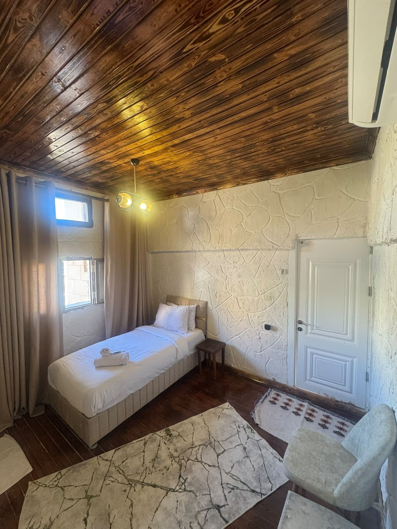 A room with a wooden ceiling, a bed with white bedding, a stone-patterned wall, a door, and a small table.