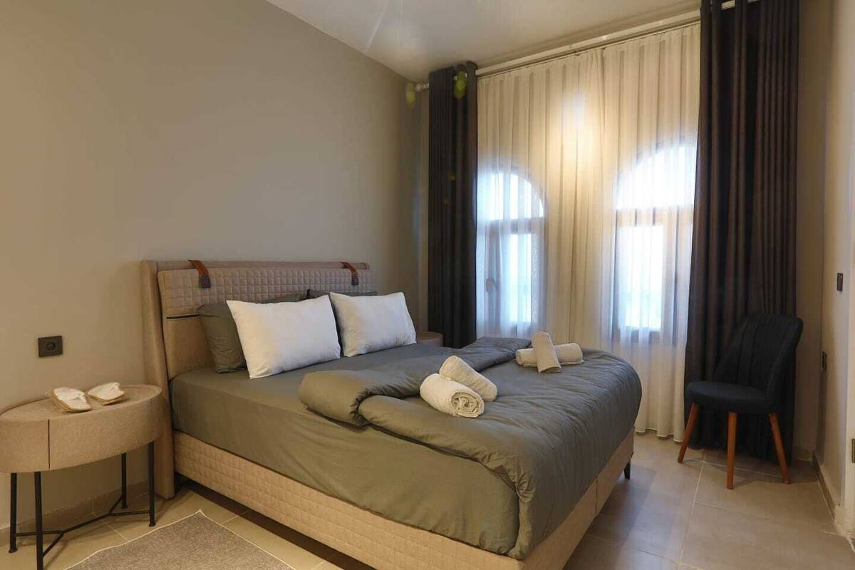 A hotel room with a bed, a chair, a nightstand, and a window with curtains.