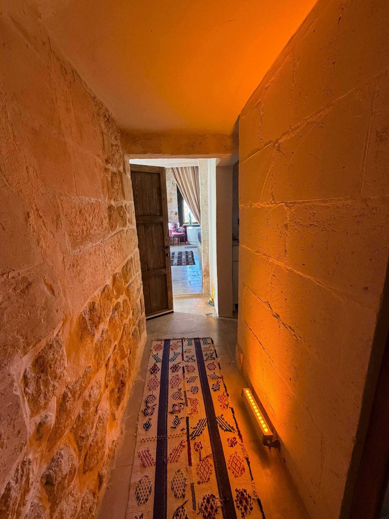 A narrow hallway with textured walls, a patterned rug, and a doorway leading to an outdoor area with a chair and table.