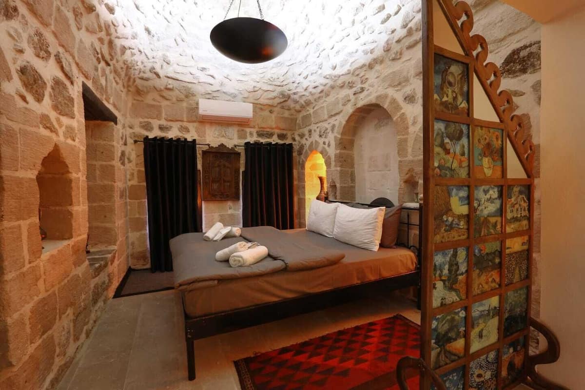 A stone-walled room with a bed, a red and black checkered rug, and a wooden panel with artwork.