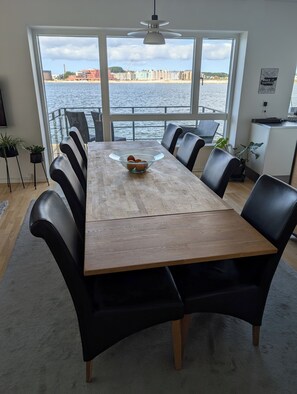 A dining area with a wooden table and black chairs, a bowl of fruit, and a view of the water and buildings. - Apartment with a breathtaking view of the Limfjord (Aalborg)