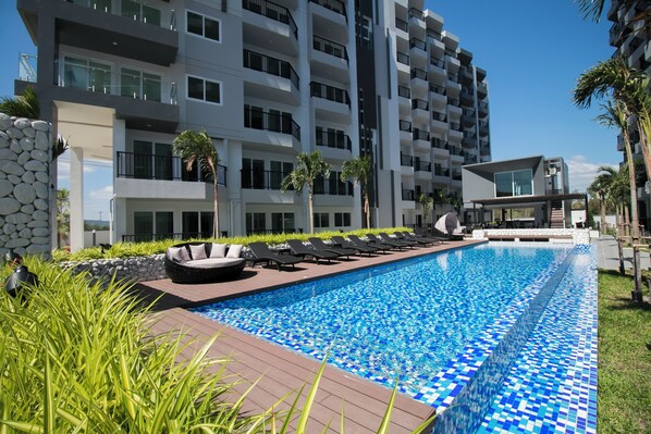A swimming pool with lounge chairs and a stone wall, surrounded by a modern building with balconies. - Mini Studio Lake view on High floor, AC, Kitchen, Balcony (Klaeng/Kram)