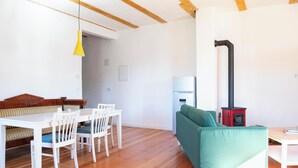Uma sala de estar moderna com piso de madeira, um sofá verde, uma mesa de jantar branca com cadeiras, uma luminária pendente amarela e uma lareira a lenha vermelha.