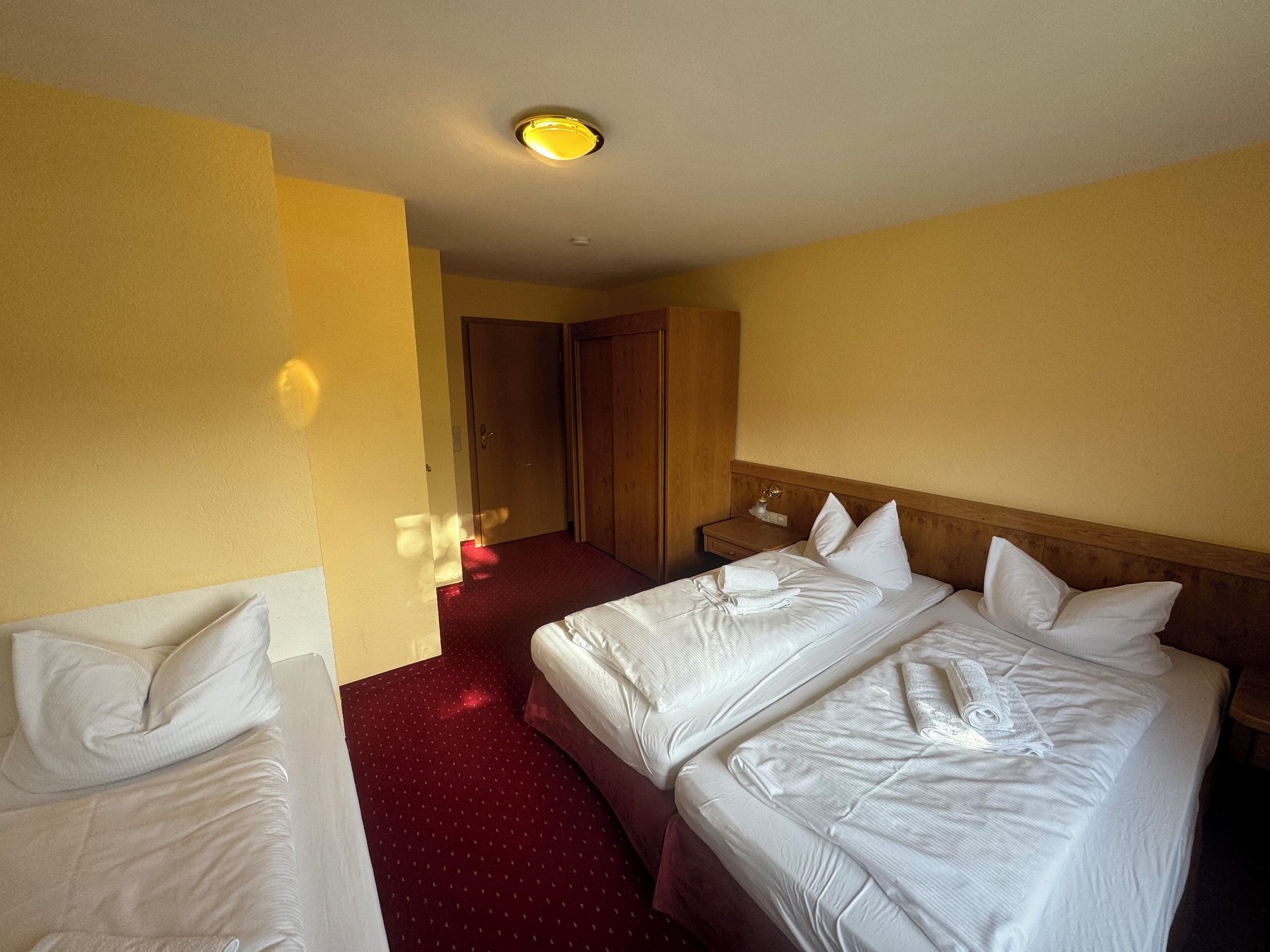 Une chambre d’hôtel avec deux lits, une tête de lit en bois, une table de chevet avec une lampe et un sol recouvert d’une moquette rouge.