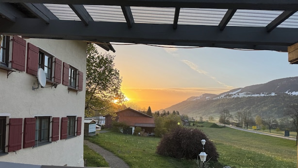 A view from a balcony overlooking a sunset, a grassy area, and a building with red shutters.