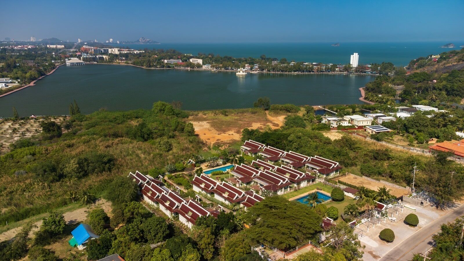 An aerial view of a resort with multiple bungalow-style buildings arranged in a grid pattern, a central swimming pool, and a large body of water in the background.