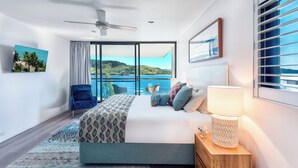 Un dormitorio moderno con una cama grande, ventilador de techo, vista al mar y balcón con sillas.