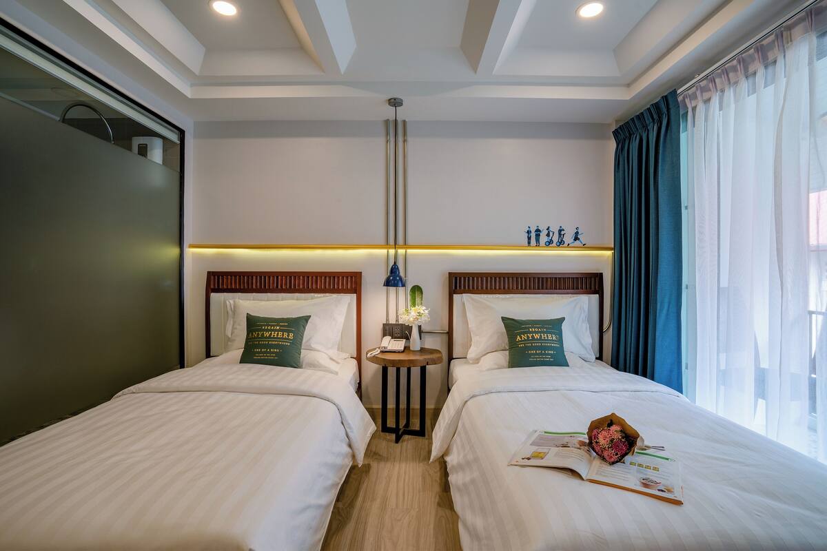 Habitación de hotel con dos camas, una mesita de noche con lámpara y vistas al exterior a través de una ventana con cortinas.