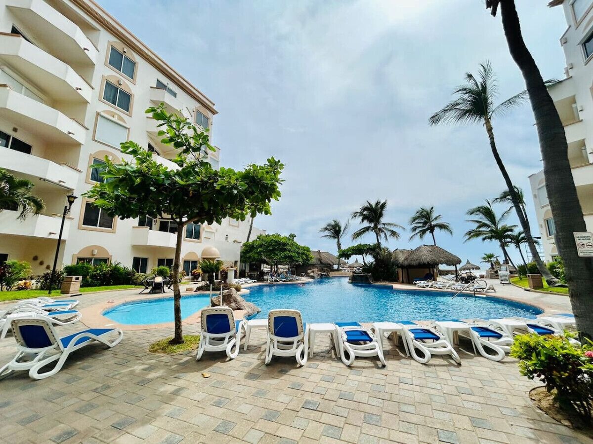 A pool area with lounge chairs, a palm tree, and a building with balconies.