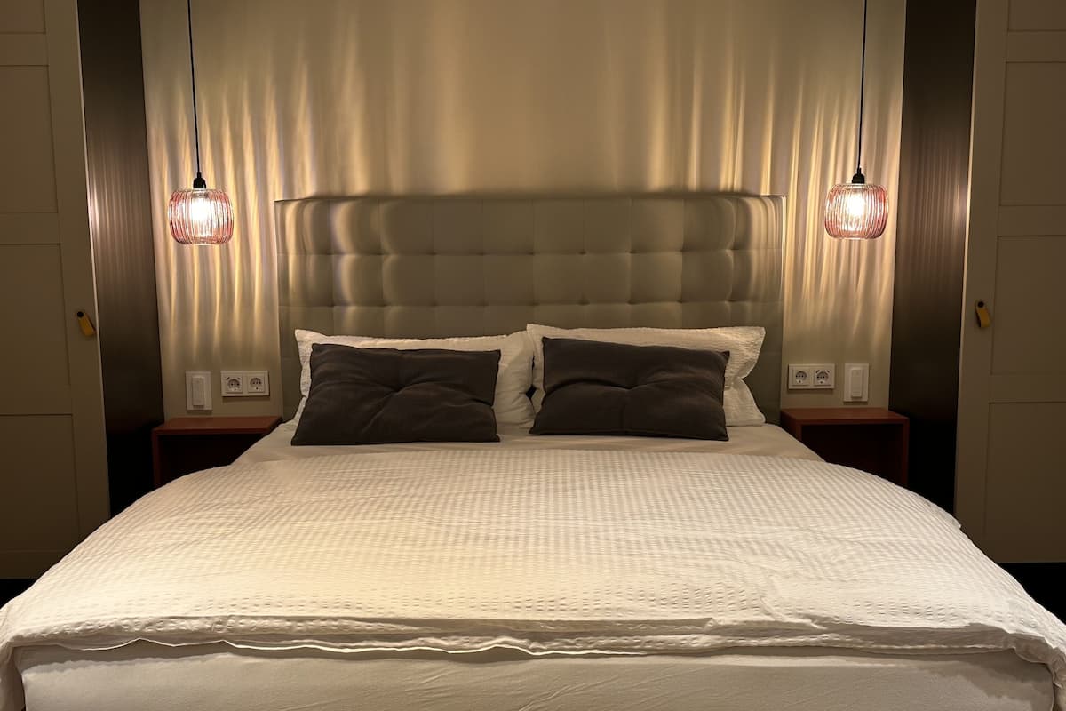 A neatly made bed with a textured white duvet, two dark pillows, and a tufted headboard. There are two bedside tables with lamps, and a power outlet on the wall.
