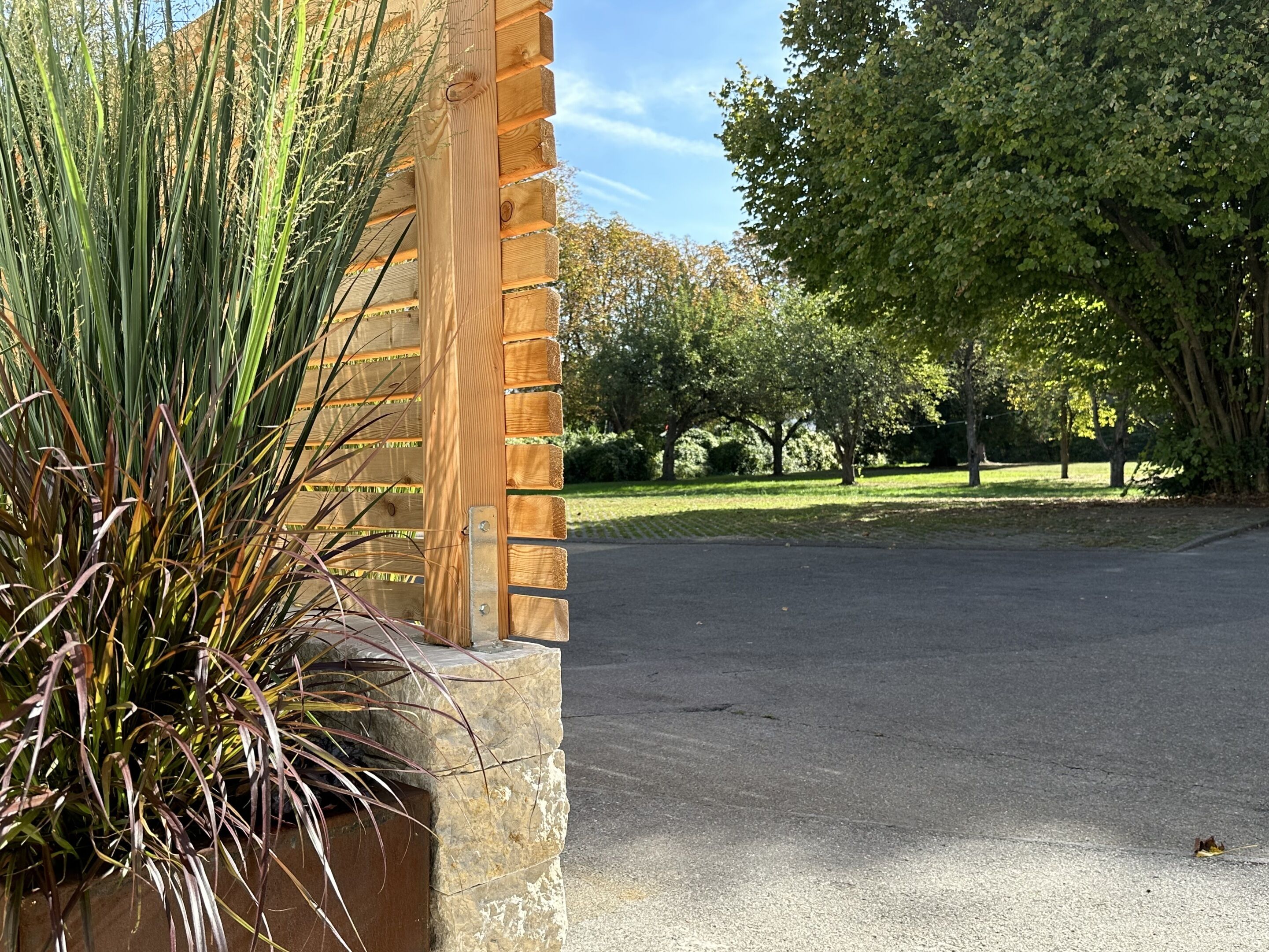 A paved driveway leading to a wooden gate with a stone base, surrounded by lush greenery and trees.