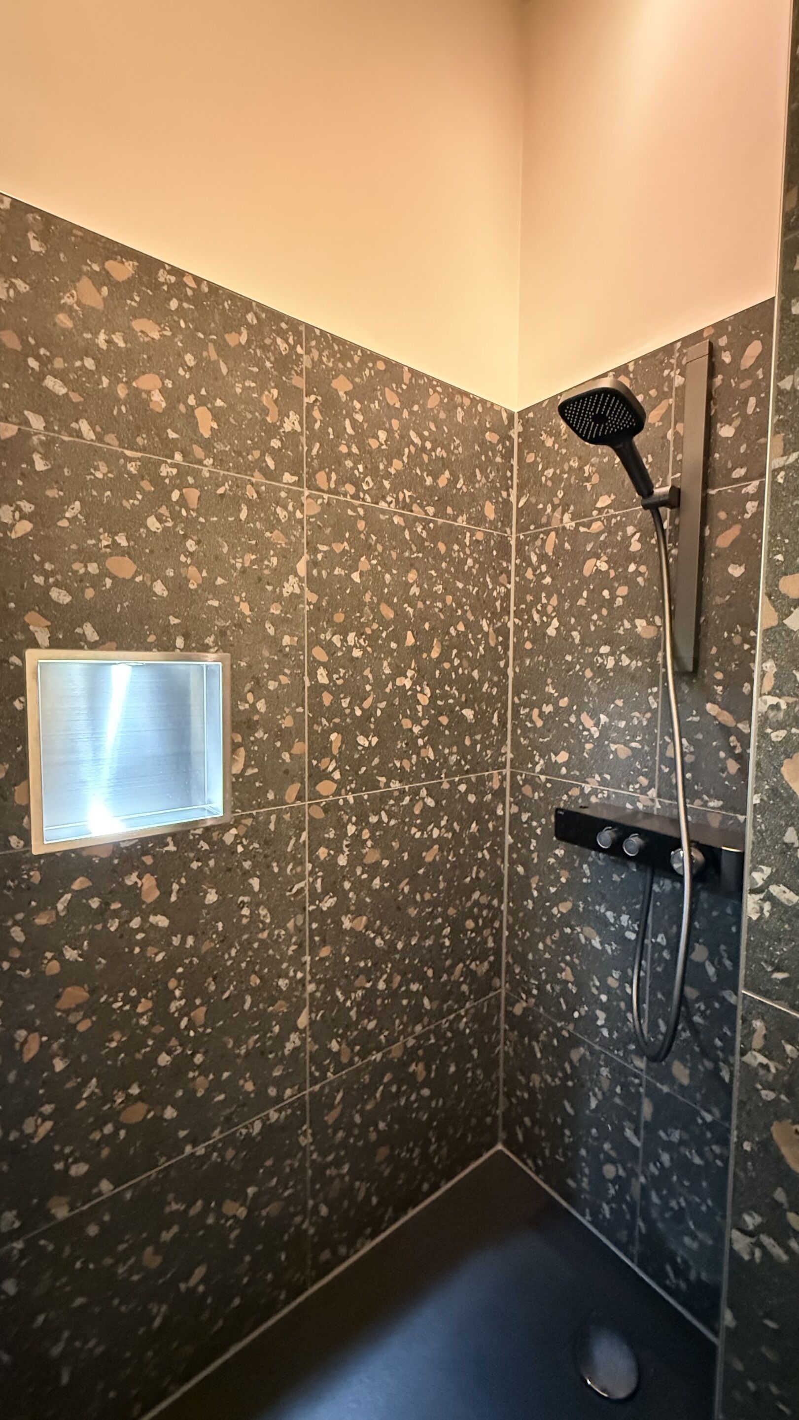A shower with a handheld showerhead, a wall-mounted control panel, and a small window.