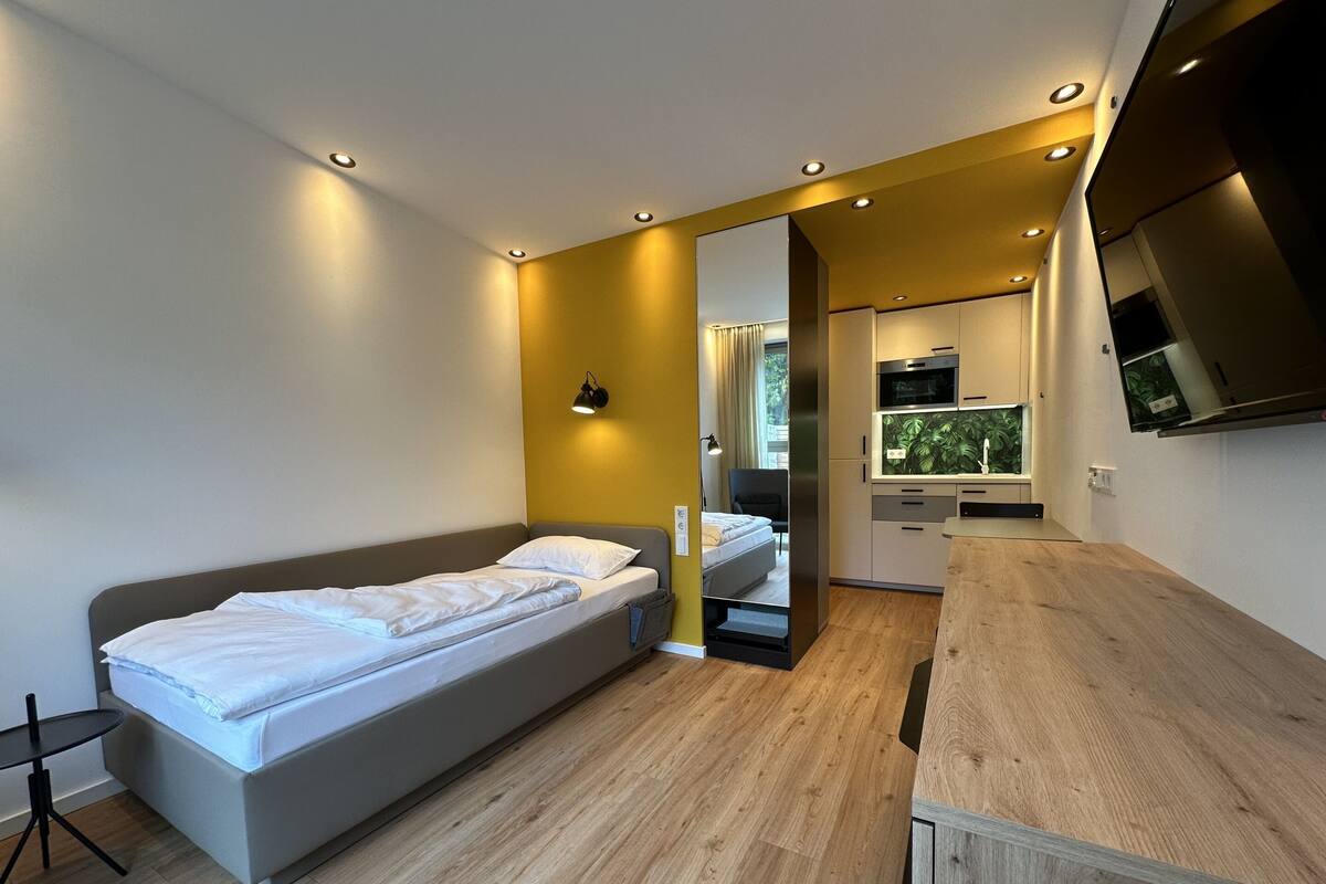 A modern bedroom with a single bed, a small desk, and a kitchenette area.