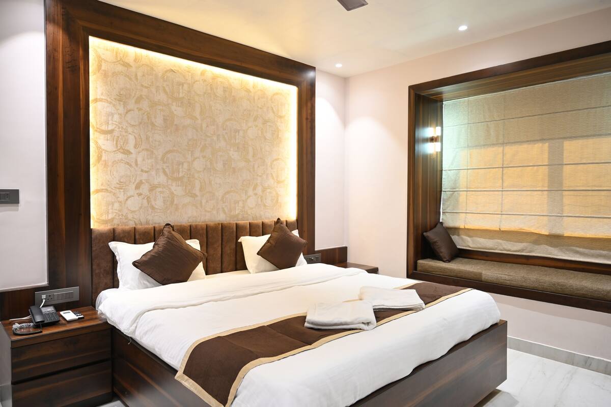 A modern hotel room with a large bed, a built-in headboard, a bedside table with a phone, and a window with blinds.